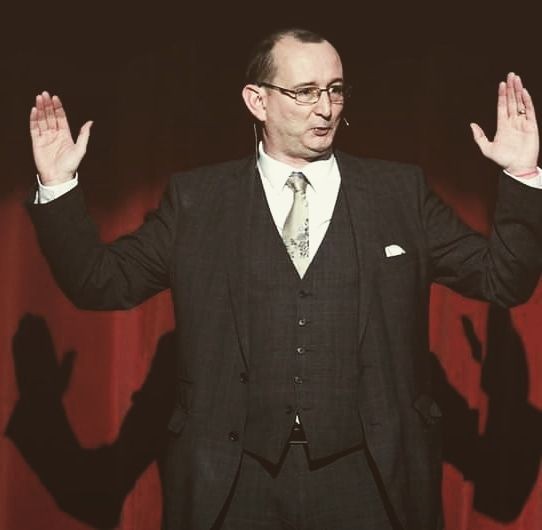 Man in a suit with hands raised, standing on stage against a red curtain background.
